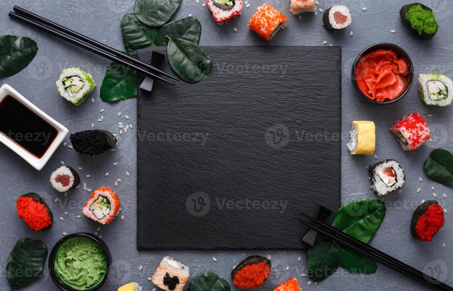 Top view on traditional japanese empty square black slate plate framed with sushi, ginger, soy, wasabi bowls, green leaves and chopsticks on rustic grey backround copy space photo
