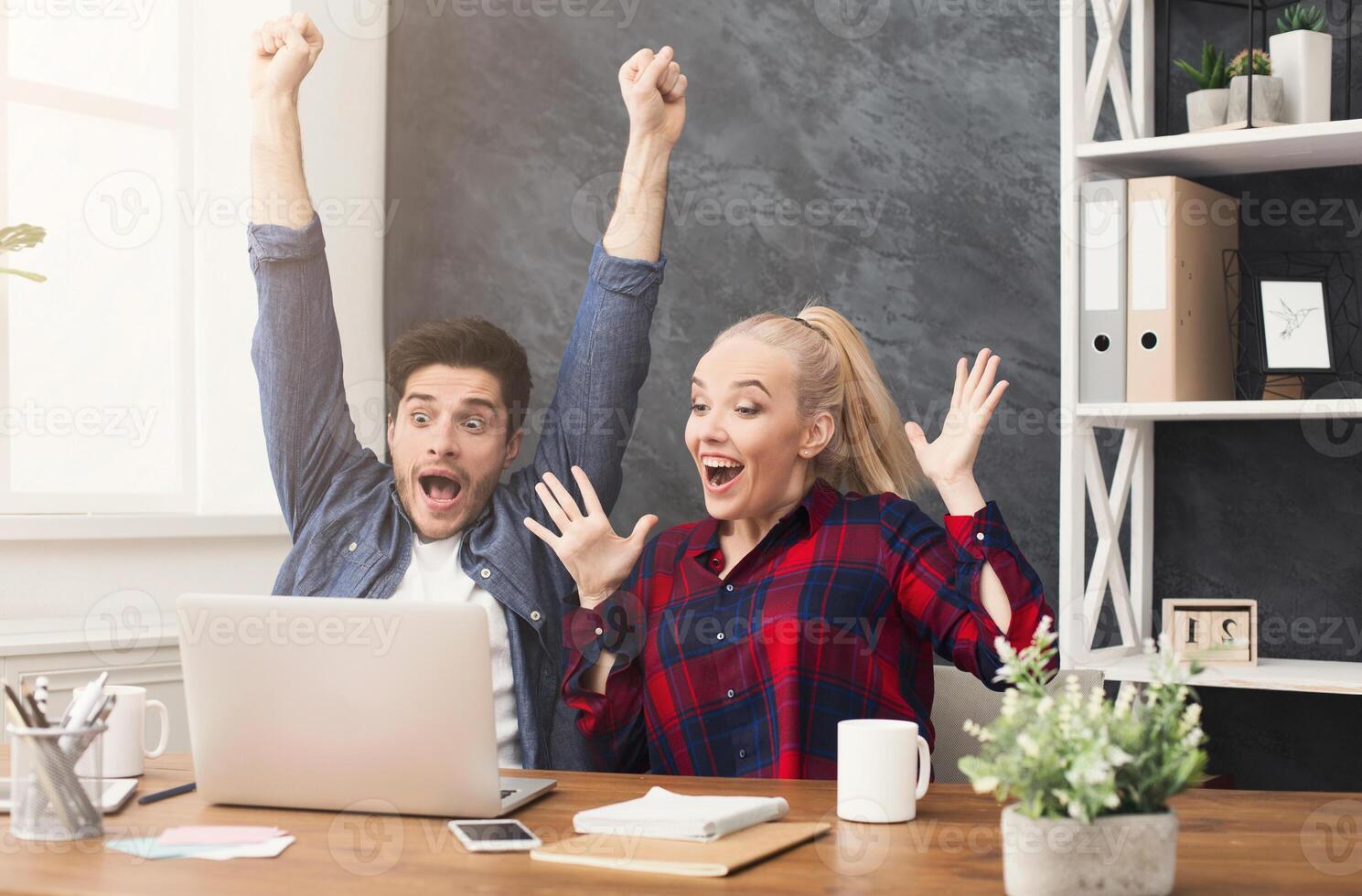 Winner business partners happy, successful, in white modern office, sitting at workplace. Colleagues raising their arms photo