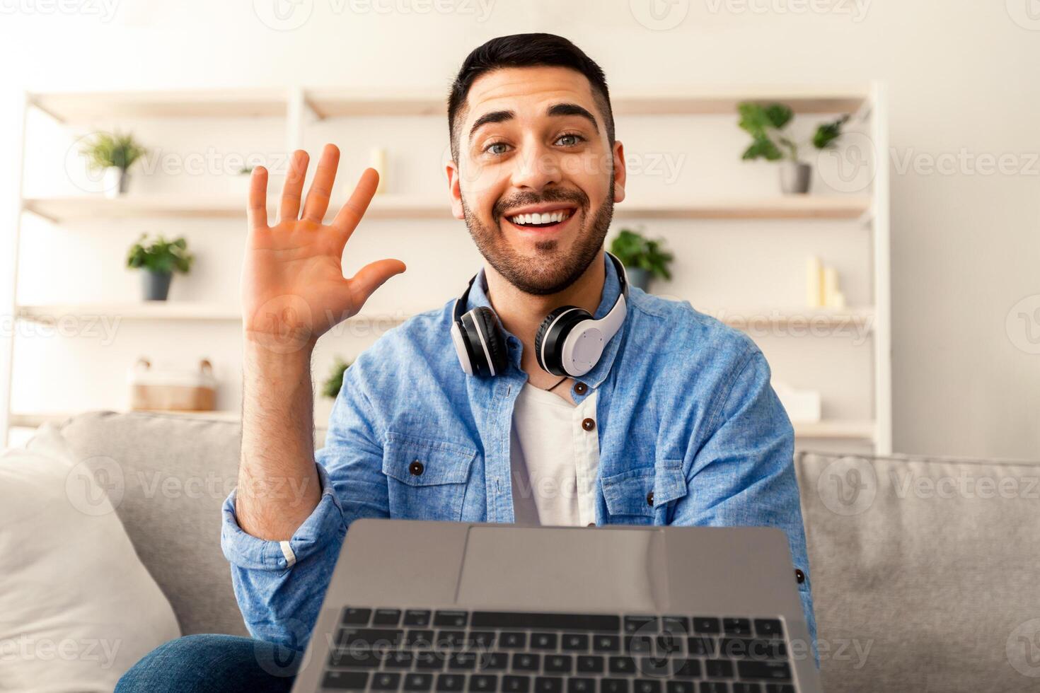 Online Call. Portrait of smiling bearded man having virtual conference with friends, family or colleagues using computer, waving hello with hand, sitting on couch at home office, webcamera view photo