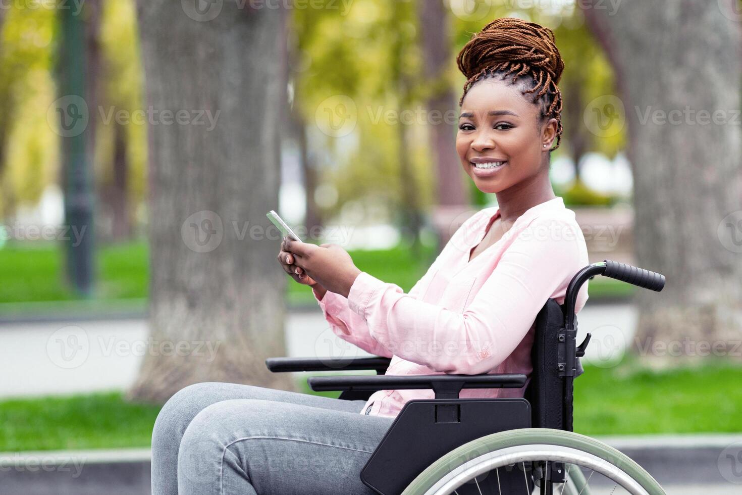 Happy disabled black woman in wheelchair using cellphone, checking social media, browsing web at autumn park. Cheerful disabled Afro lady with mobile device watching , chatting to friend outdoors photo
