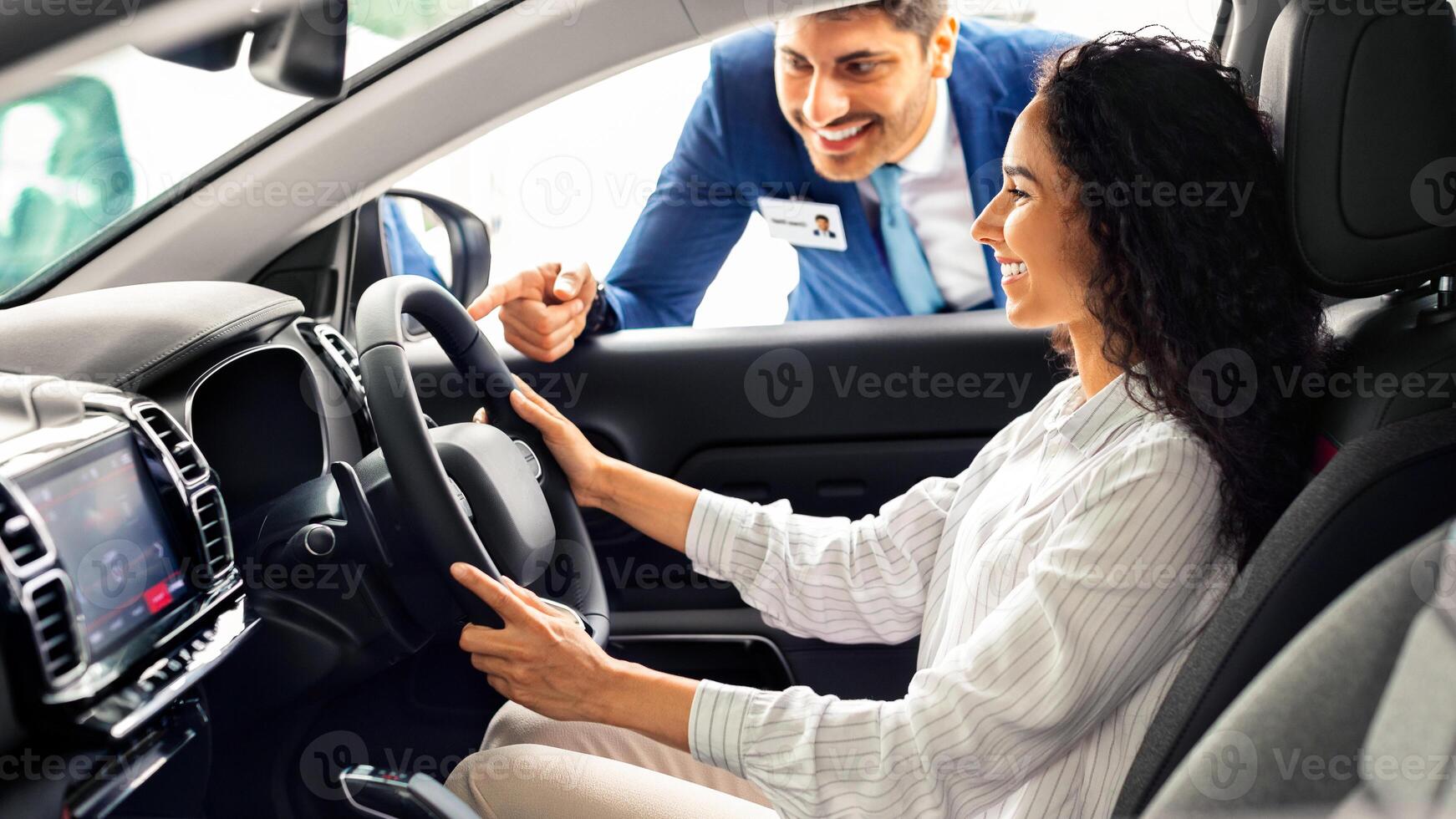 Young business lady buying new luxury car in new showroom, happy brunette woman sitting in nice automobile, holding hands in steering wheel, having conversation with sales assistant, copy space photo