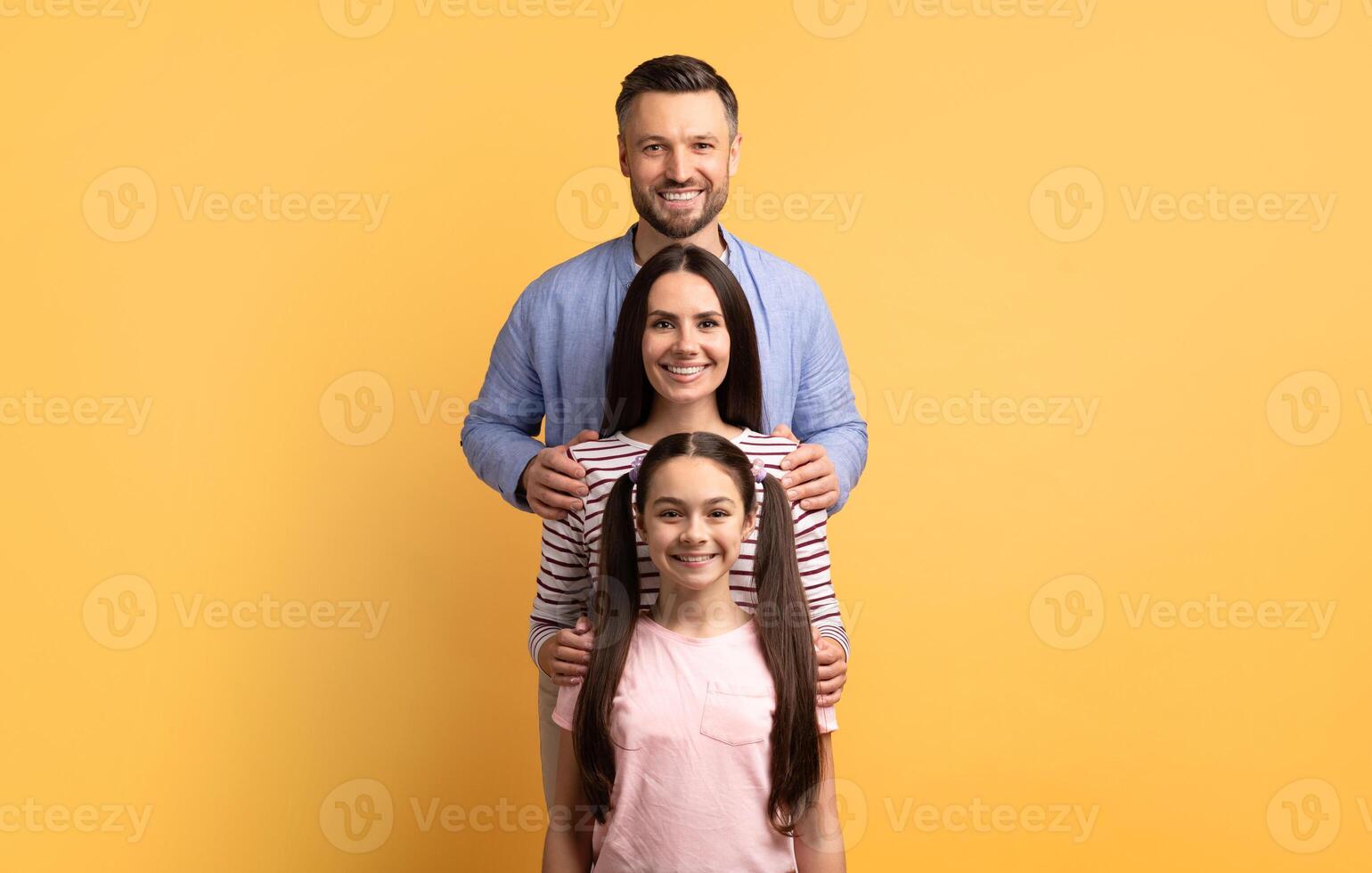 A family of three is standing together against a bright yellow background. The father, mother, and daughter show love and affection, smiling happily and bonding in this joyful moment. photo