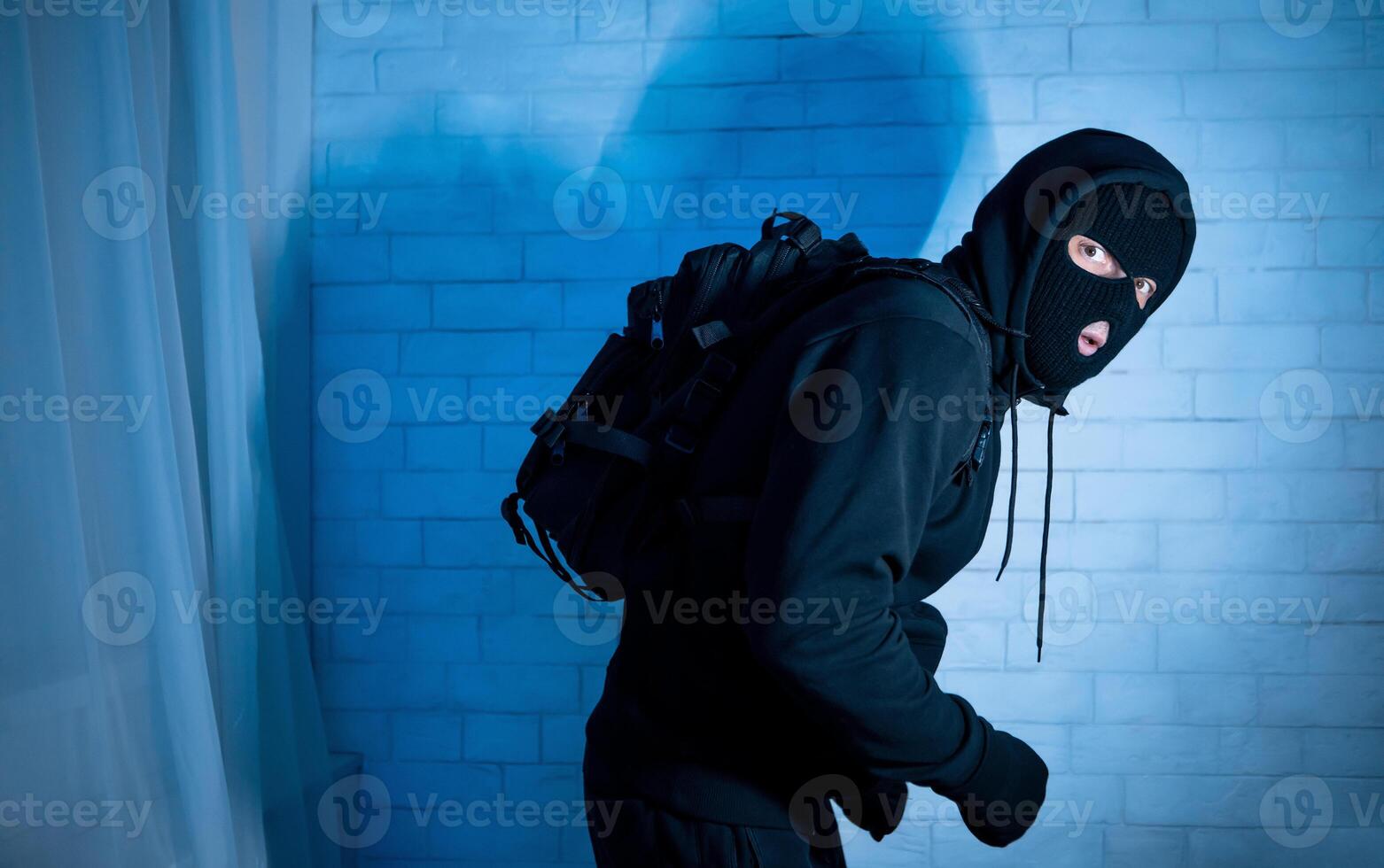 Burglary Concept. Sneaky and scared intruder wearing black balaclava hat lurking in the dark, looking at camera photo