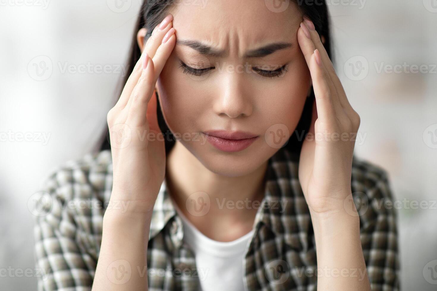 Closeup of young asian woman having headache, touching her temples, home interior. Brunette lady feeling upset or suffering from migraine, sitting on couch in living room with eyes closed photo