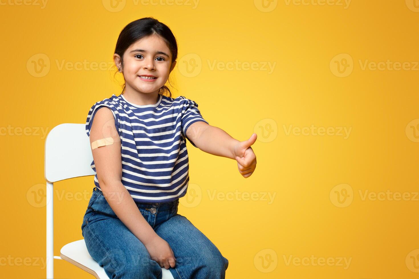 Kids Vaccination. Vaccinated Little Arab Girl Gesturing Thumb Up After Covid-19 Vaccine Injection, Cheerful Middle Eastern Child With Adhesive Band On Arm Sitting On Chair Over Yellow Background photo