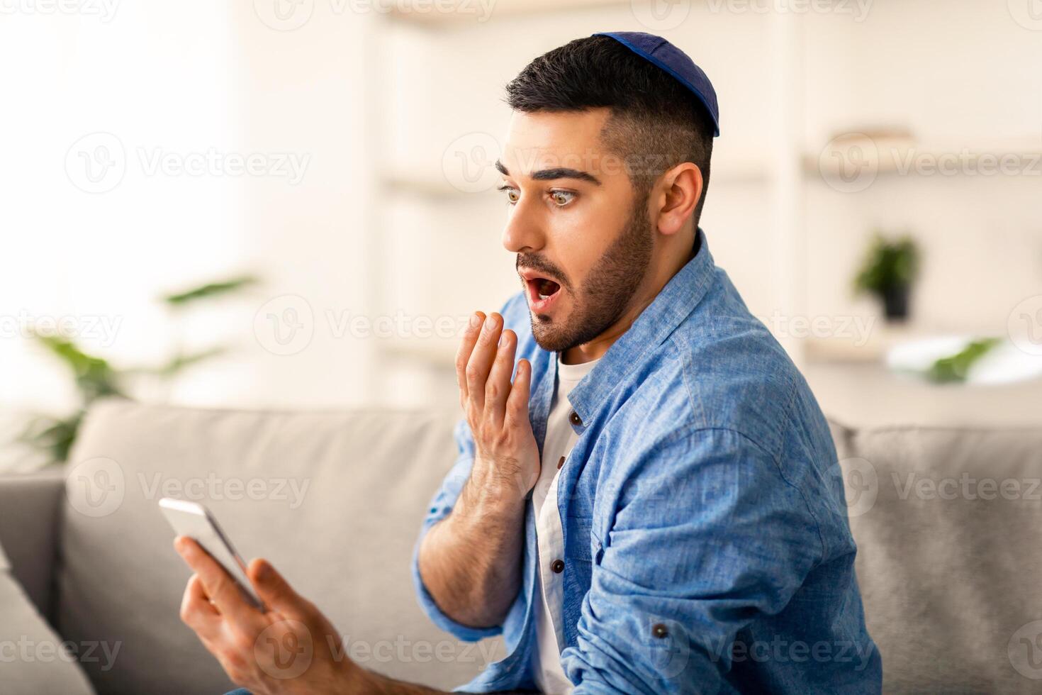 Oh No. Portrait of shocked frustrated jewish man having problems, holding and looking at cellphone screen sitting on couch at home, surprised guy in kippa closing covering mouth with hand, free space photo