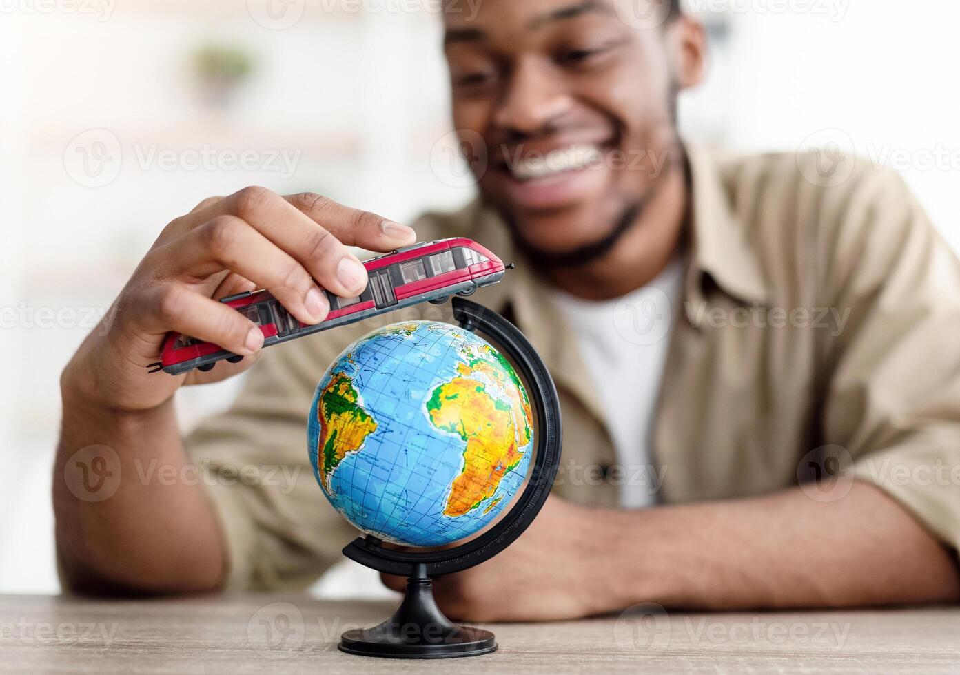 Travel Concept. African American Man Playing With Train Model And World Globe Sitting Indoors. Guy Dreaming About Trip Planning Summer Vacation. Selective Focus On Toy, Shallow Depth photo