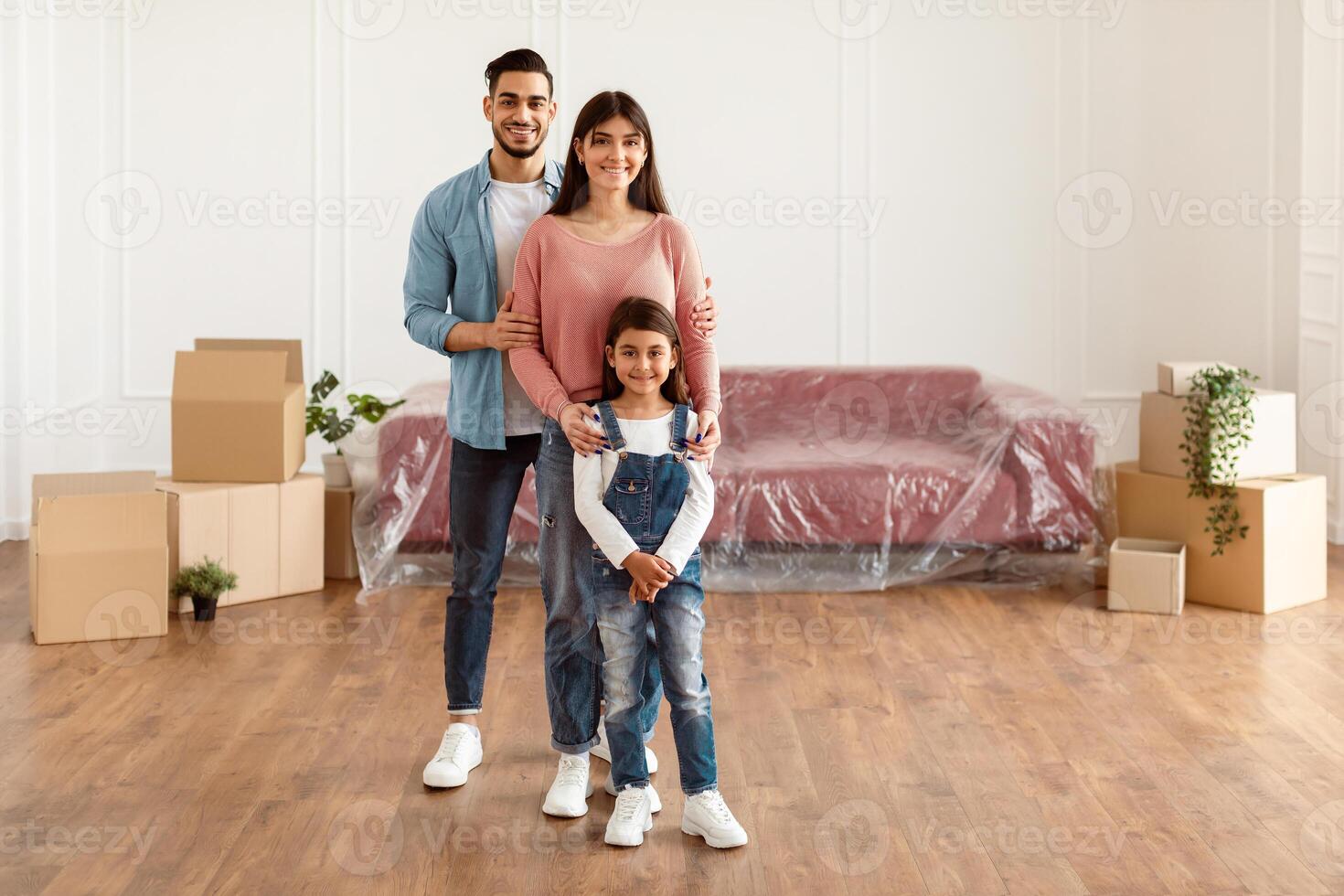 House Move, Renovation And Repair Concept. Full body length of cheerful young family of three people standing in new empty apartment, posing and looking at camera, hugging, free copy space photo