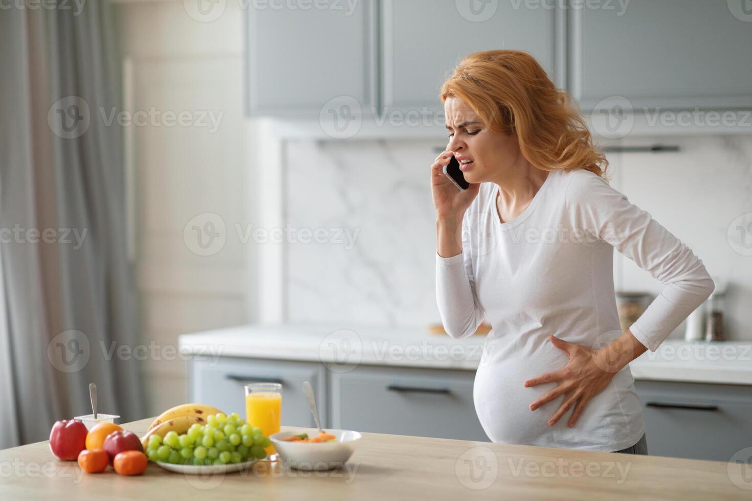 A pregnant woman stands in her kitchen, one hand on her belly while talking on the phone with a concerned expression. Fresh fruits are on the counter, highlighting her home environment, copy space photo
