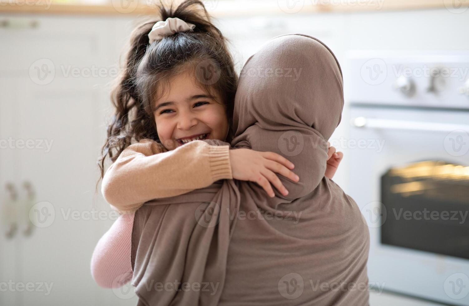 Cute Little Girl Hugging Her Muslim Mom In Headscarf And Smiling, Loving Family Bonding With Each Other In Kitchen At Home, Caring Islamic Mother Embracing Her Laughing Child, Copy Space photo