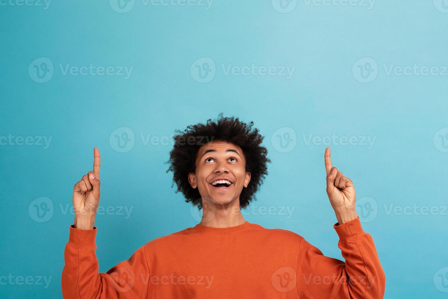 A young African American man with curly hair wears an orange sweater and smiles widely while pointing upward at copy space with both hands. The vibrant blue background enhances his joyful expression. photo