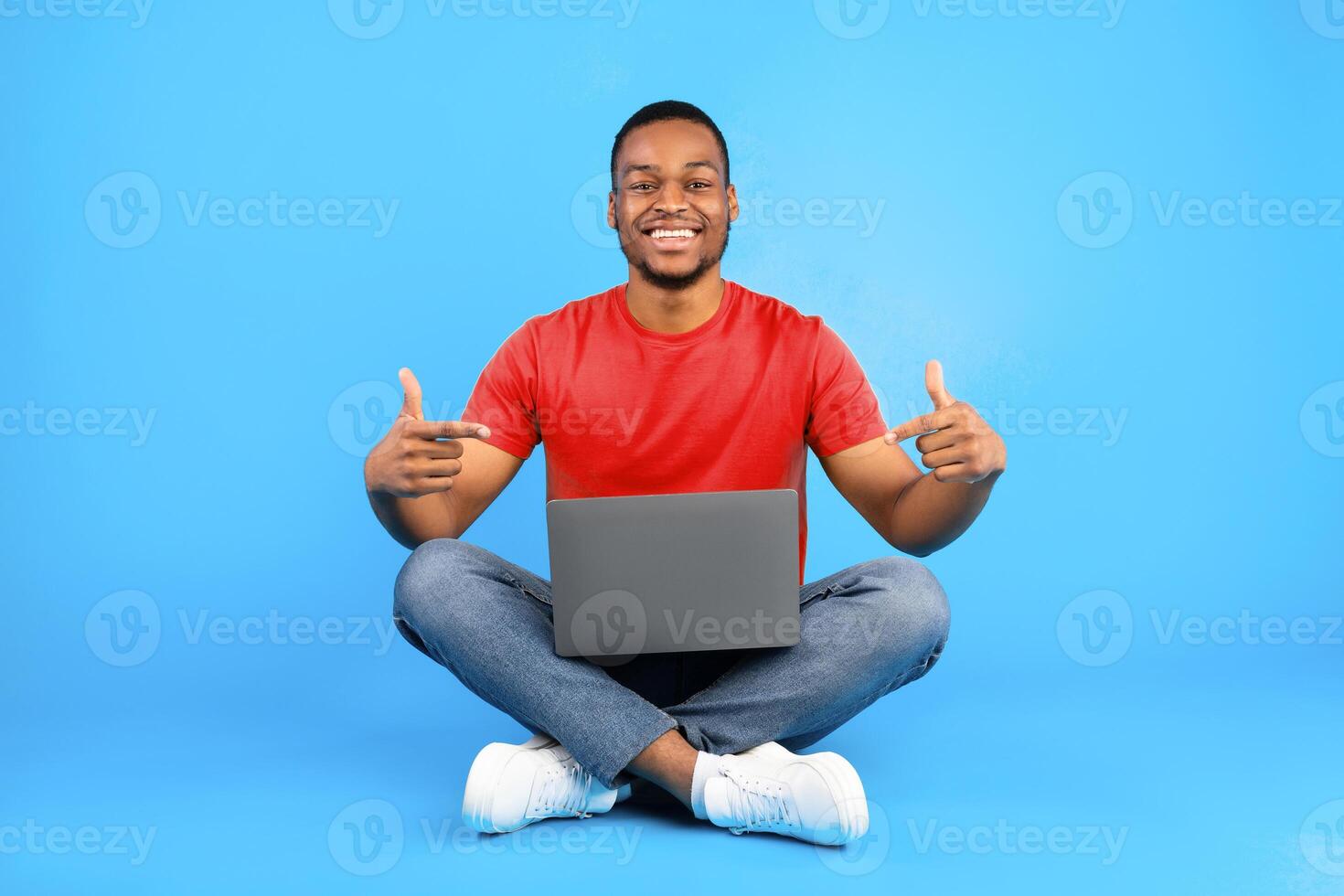 Successful Black Male Freelancer Pointing Fingers At Laptop Smiling To Camera Sitting Over Blue Background. Studio Shot Of Happy Millennial Guy Posing With Computer. Technology Concept photo