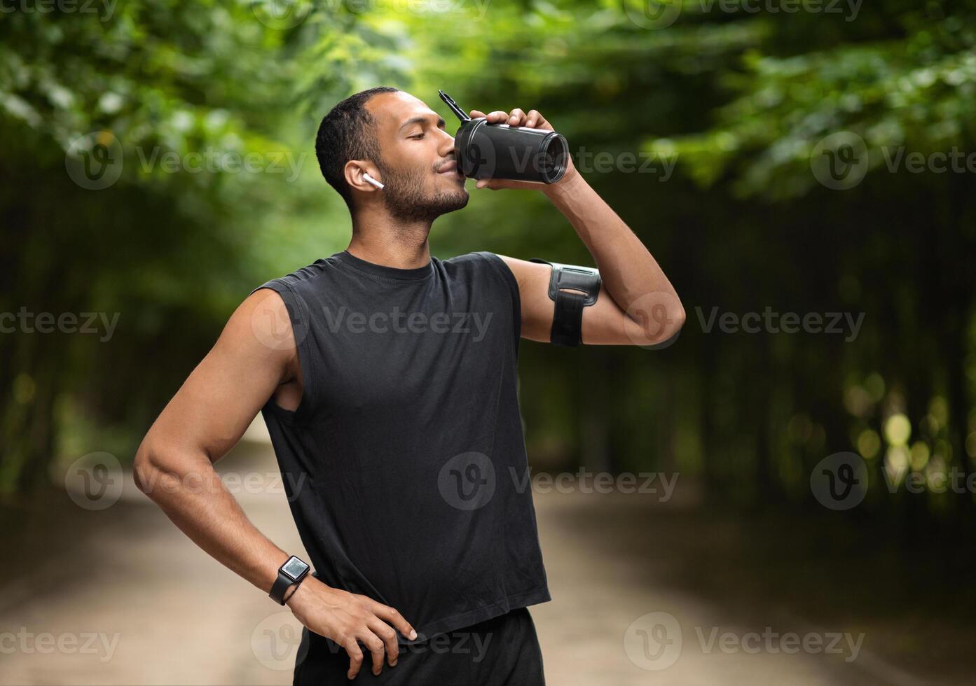 Sporty young black man drinking protein cocktail, training at public park, using wireless earbuds, listening to music while jogging by forest, copy space. Sports nutrition concept photo