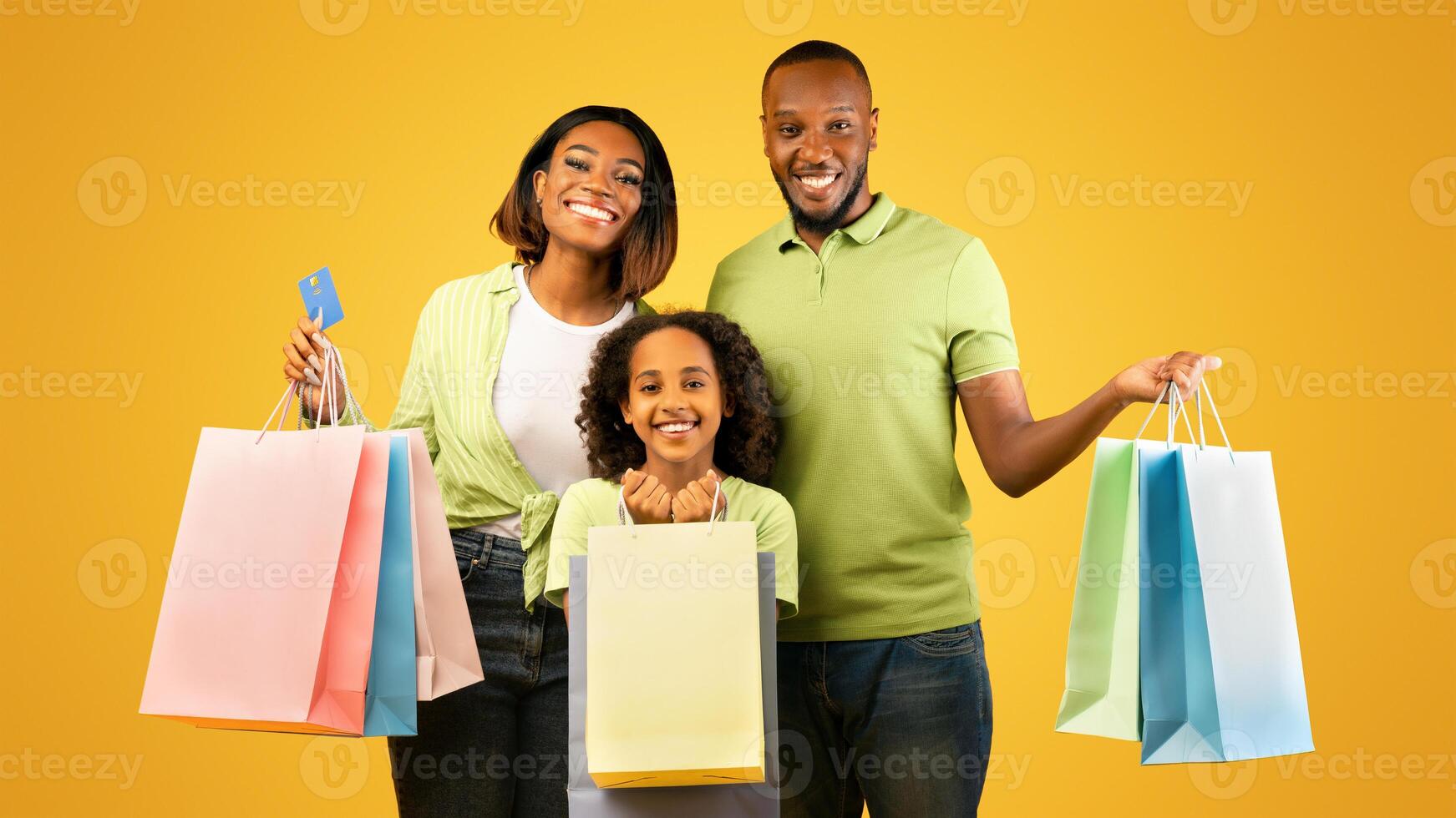 Family shopping. Excited african american parents and their cute daughter holding shopper bags and smiling while posing over yellow background, woman holding credit card in hand photo
