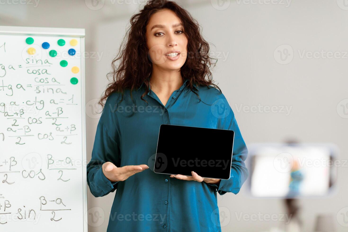 Modern Distance Learning. Lady math teacher making conference chat with students using cellphone on tripod, holding and showing tablet with empty screen with copy space for mock up, explaining photo