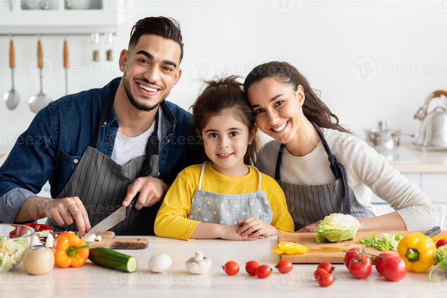 Portrait Of Beautiful Happy Middle Eastern Family With Little Daughter Cooking In Kitchen, Cheerful Arabic Parents And Cute Female Child Smiling At Camera While Preparing Food At Home Together photo