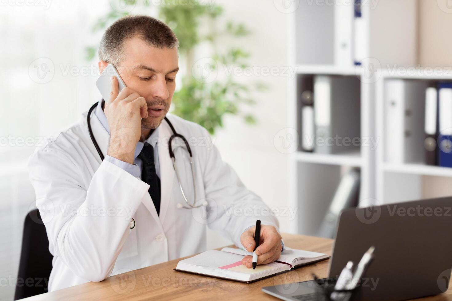General Practitioner Concept. Portrait of middle aged doctor physician talking on mobile phone with his patient and writing in his diary making notes or appointment, consulting client. Telemedicine photo