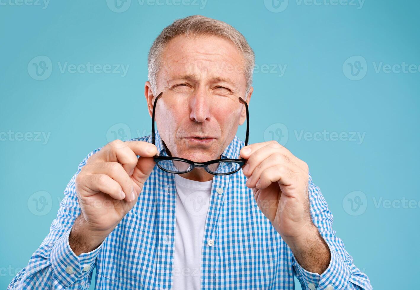 Poor Eyesight. Mature Man Can't See, Squinting Eyes Putting On Glasses Having Problems With Vision, Looking At Camera At Blue Studio. Ophtalmic Issue, Bad Sight In Older Age, Macular Degeneration photo