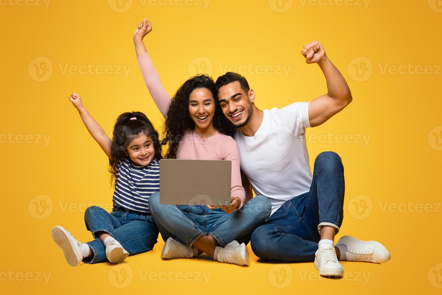 Online Win. Excited Arab Parents And Little Daughter Celebrating Success With Laptop, Happy Middle Eastern Family Exclaiming With Joy While Looking At Computer Screen, Sitting On Yellow Background photo