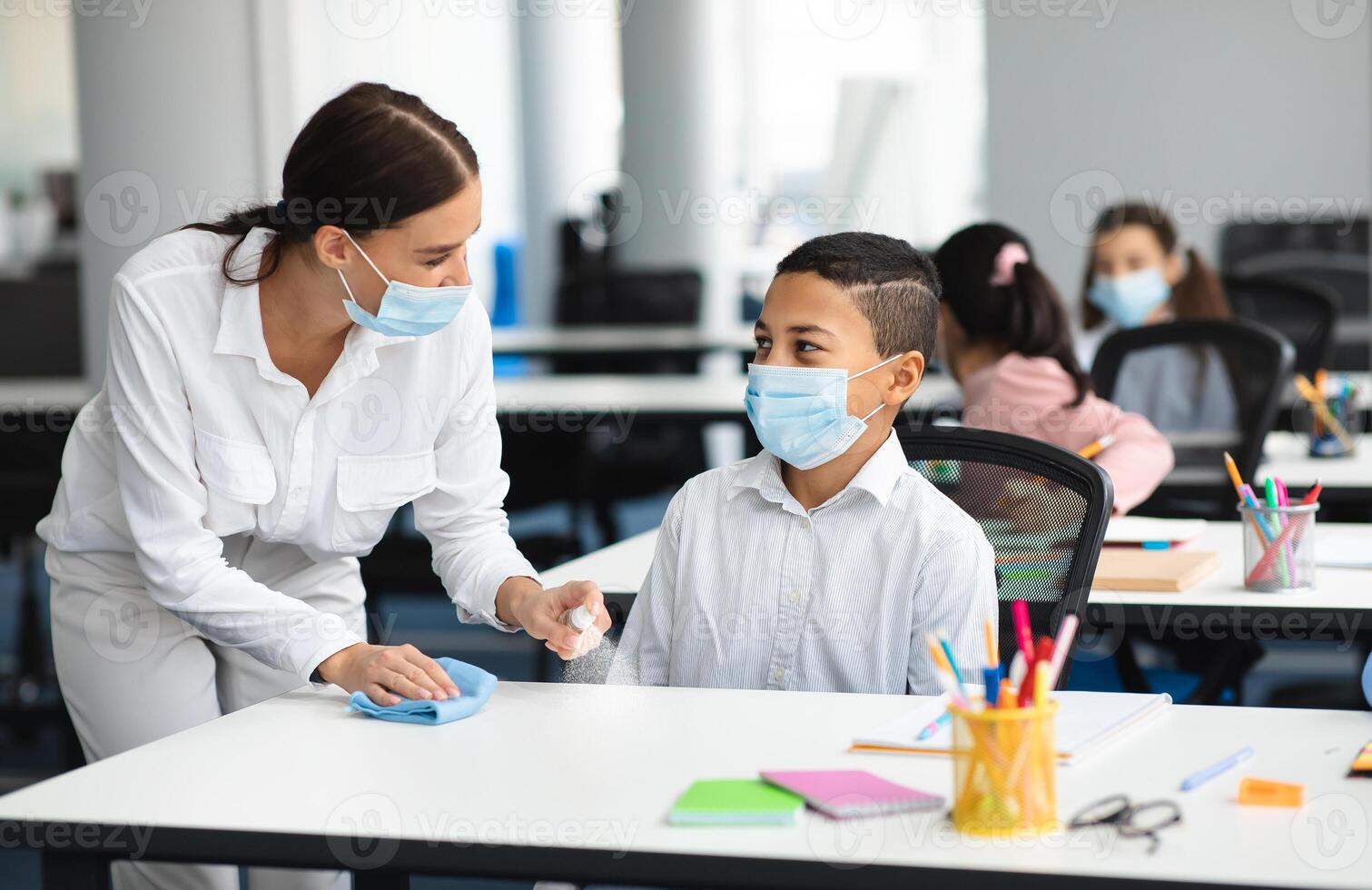 Hygiene And Cleaning. Teacher cleaning table with antibacterial sanitizer and napkin, disinfecting surface, applying antiseptic spray on desk, wearing disposable face mask, boy studying in classroom photo