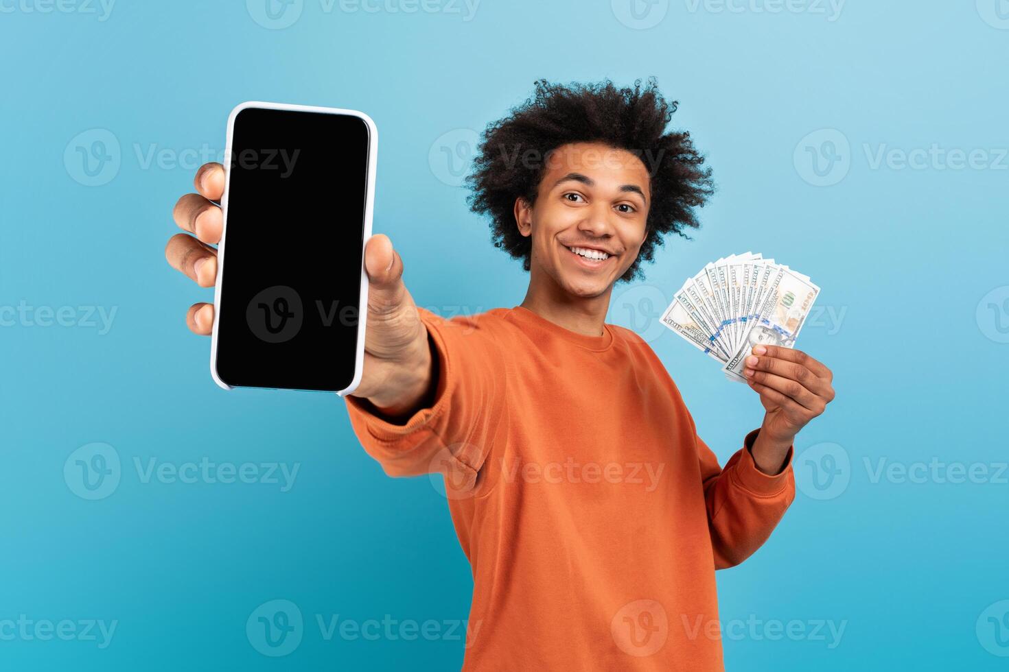 A smiling African American man holds a smartphone in one hand and stacks of cash in the other. He stands against a blue background, celebrating a successful moment in life. photo