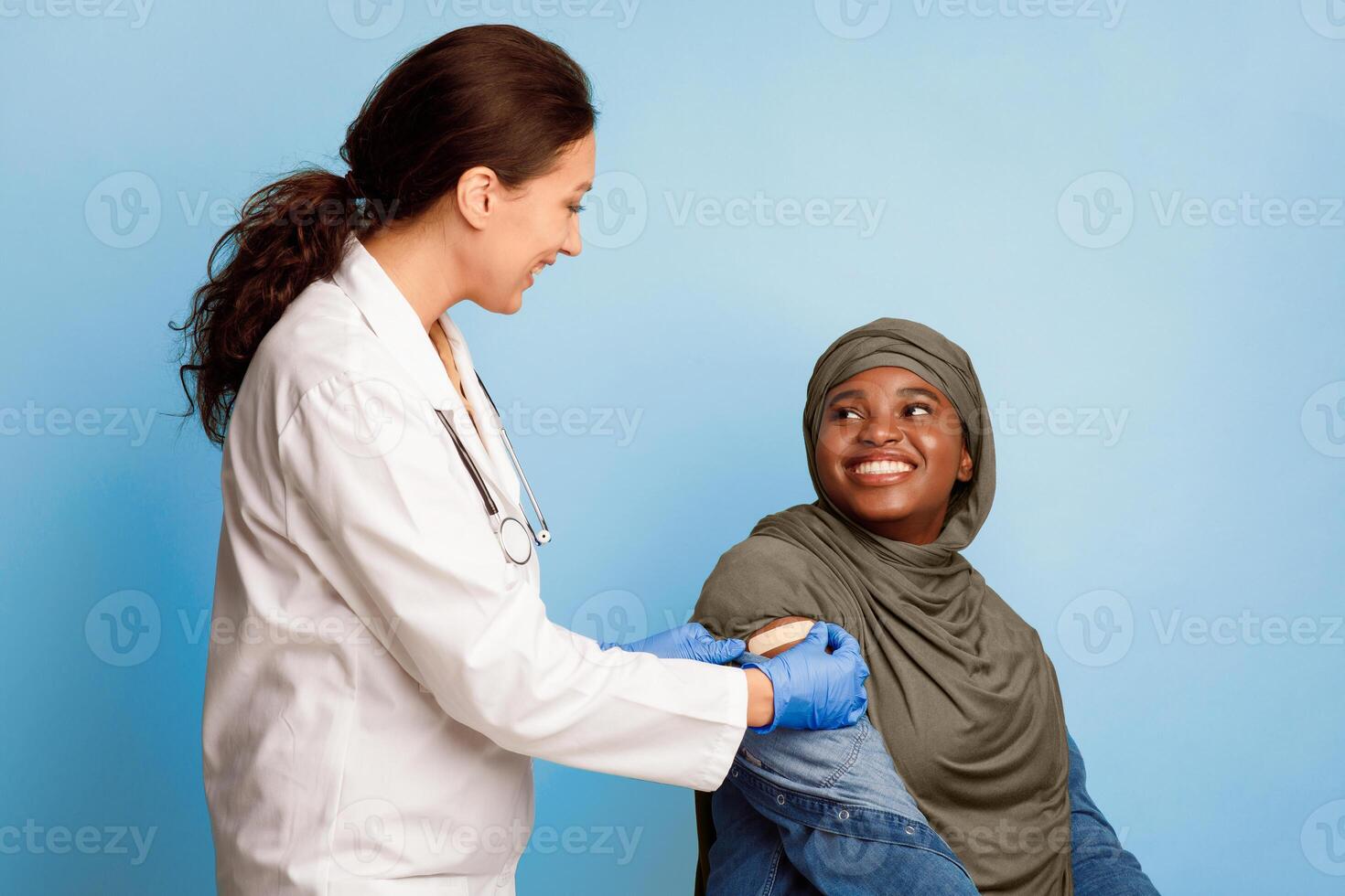 Covid-19 Vaccination. Black Muslim Female Patient Getting Vaccinated Against Coronavirus, Nurse Applying Adhesive Bandage Plaster On Vaccinated Arm After Vaccine Injection On Blue Studio Background photo