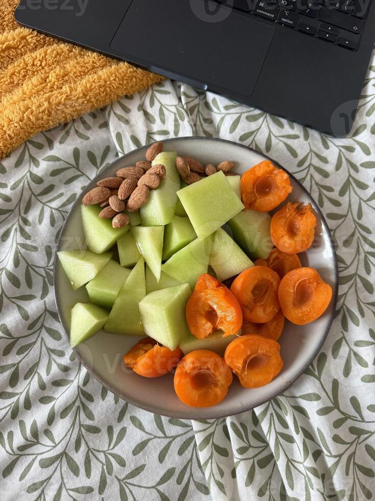 Fresh fruit platter with melon and apricots on cozy bedspread near laptop photo