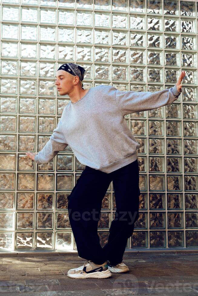 A young man in a gray sweatshirt and black pants dances in front of a glass block wall photo