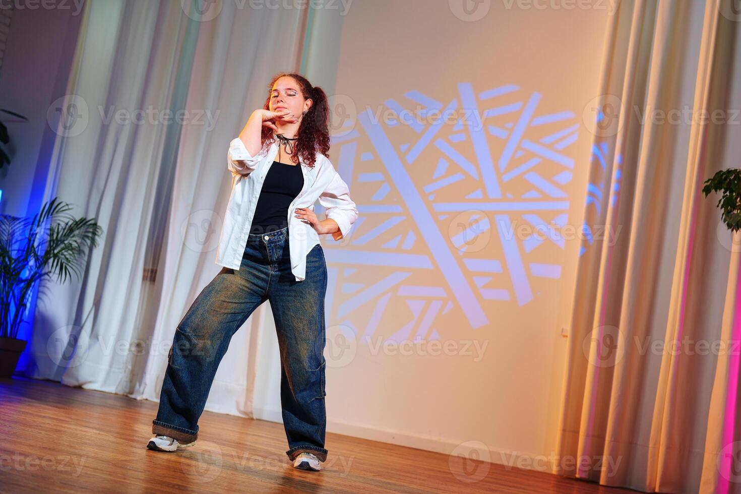A young woman in jeans and a white shirt dances on a stage with a blue light shining on her photo