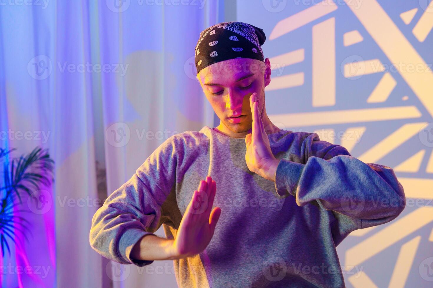 A young man with a bandana practices his dance moves in a studio with colorful lighting photo