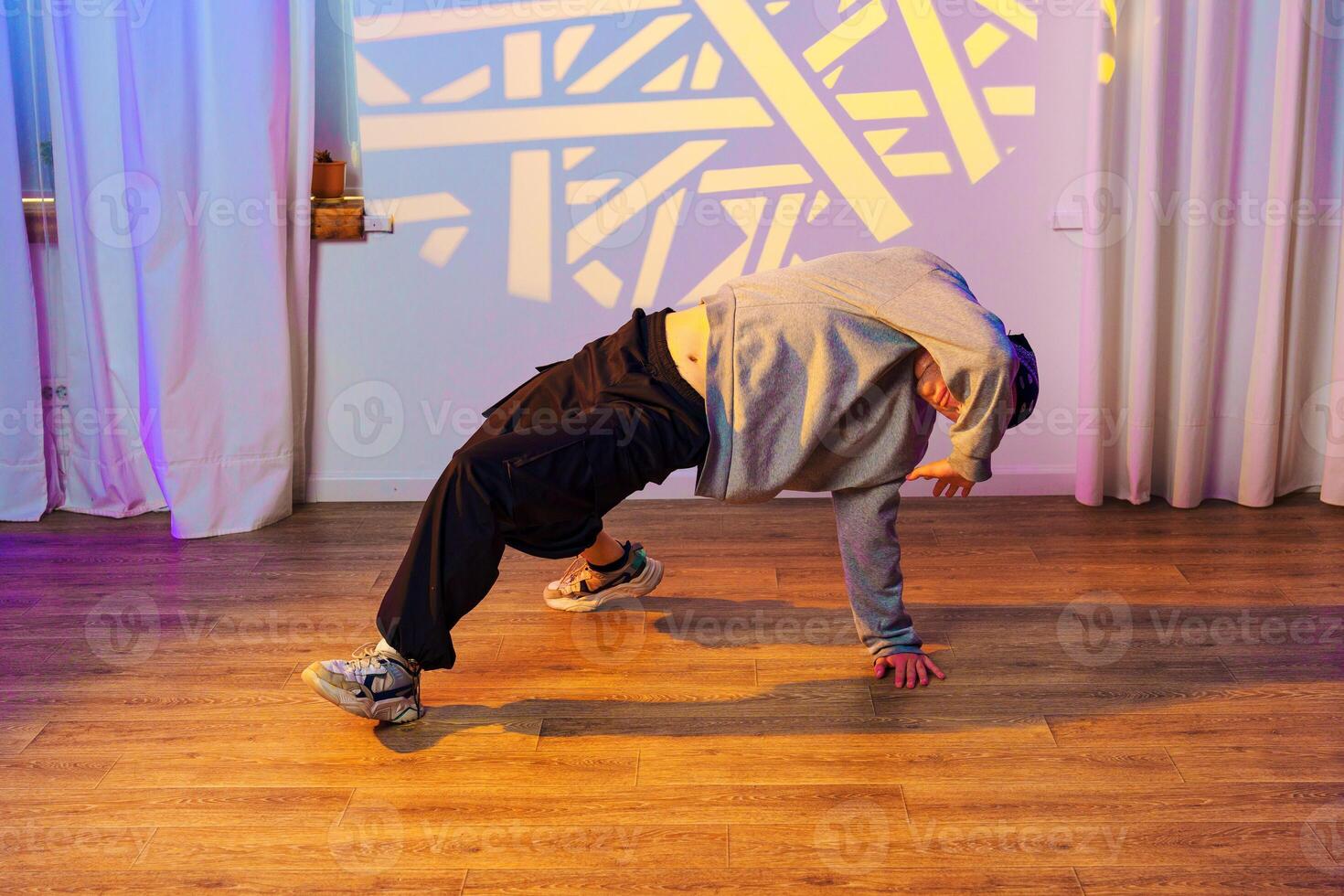 A dancer performs a breakdancing move in a studio with white curtains and a patterned light photo