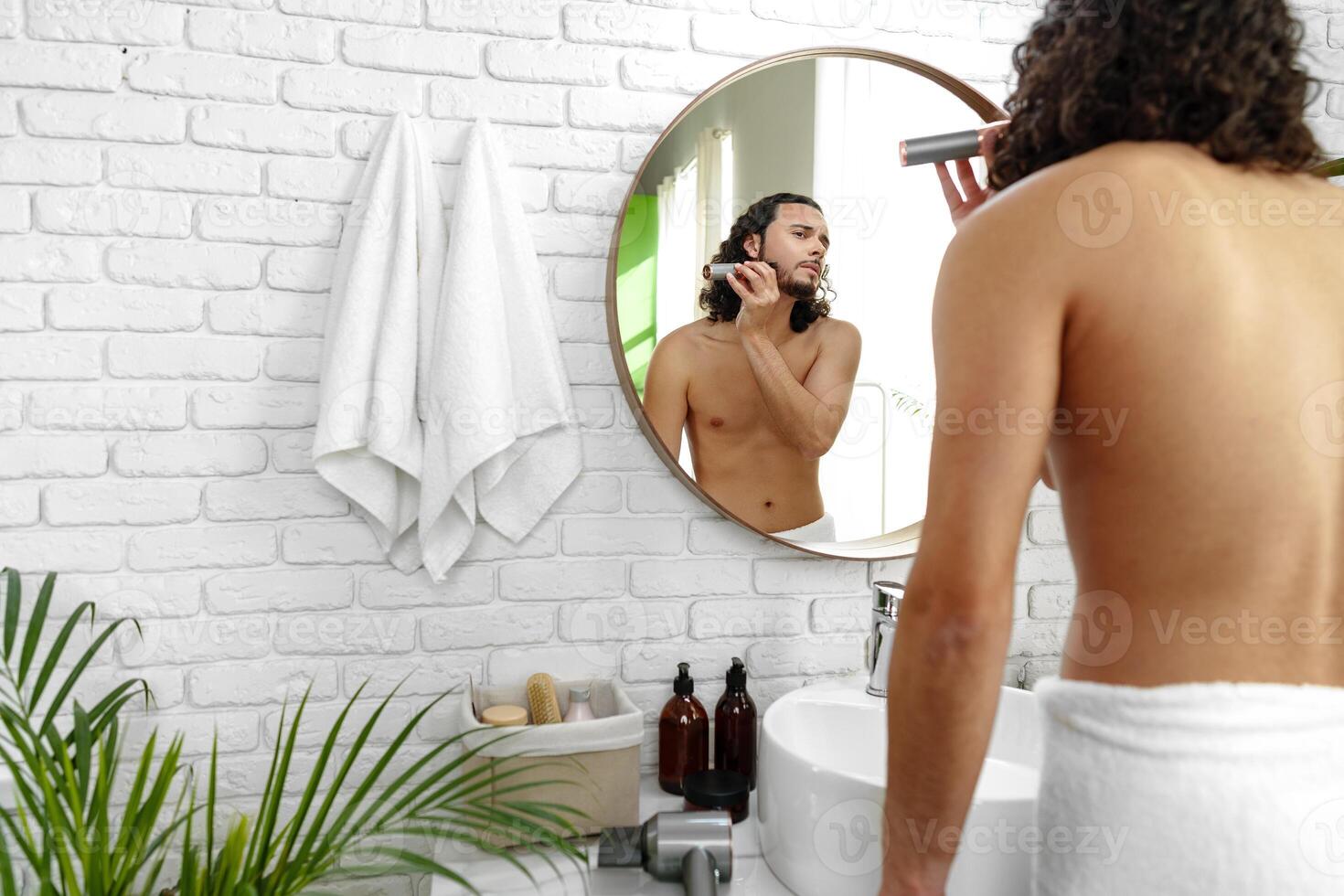 A man with curly hair grooming himself in the bathroom while looking into a round mirror photo