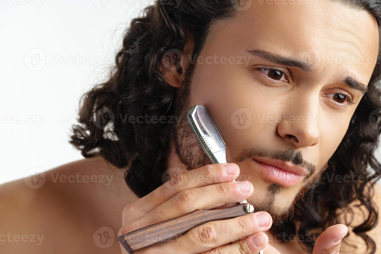 Man with curly hair grooming his beard carefully using a straight razor against a plain white background photo