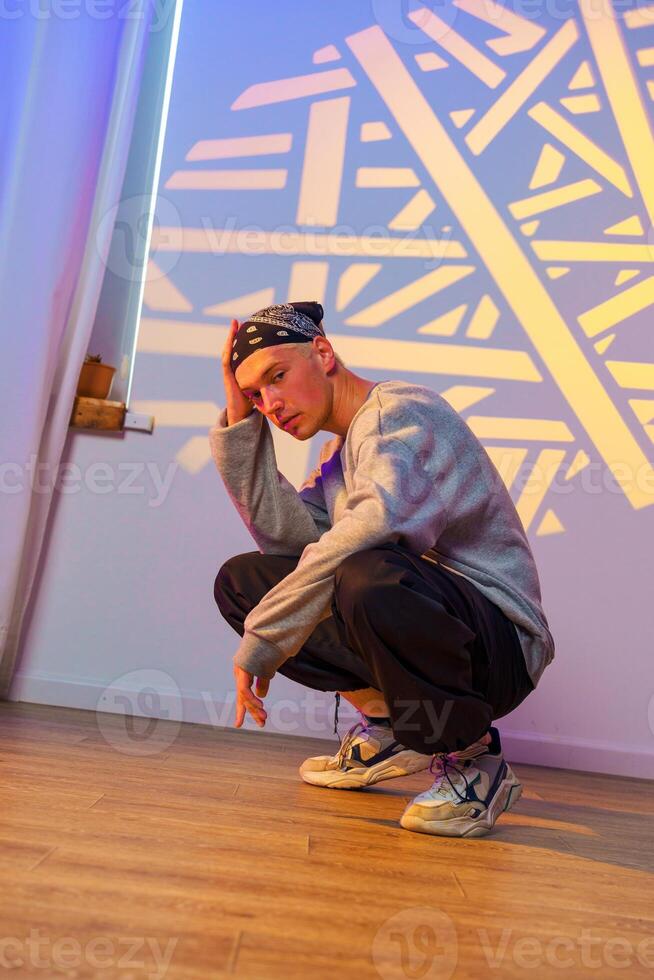A young man in a bandana and grey sweatshirt squats on a wooden floor in front of a wall with a geometric light projection photo