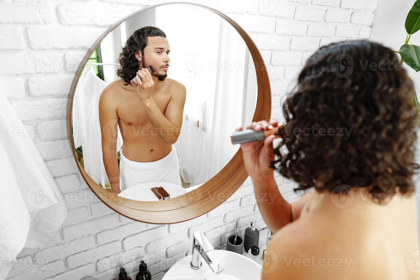 A man with curly hair grooming himself in the bathroom while looking into a round mirror photo