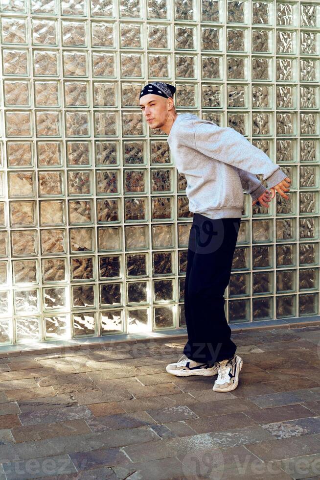 A young man in a gray sweatshirt and black pants stands in front of a glass block wall photo