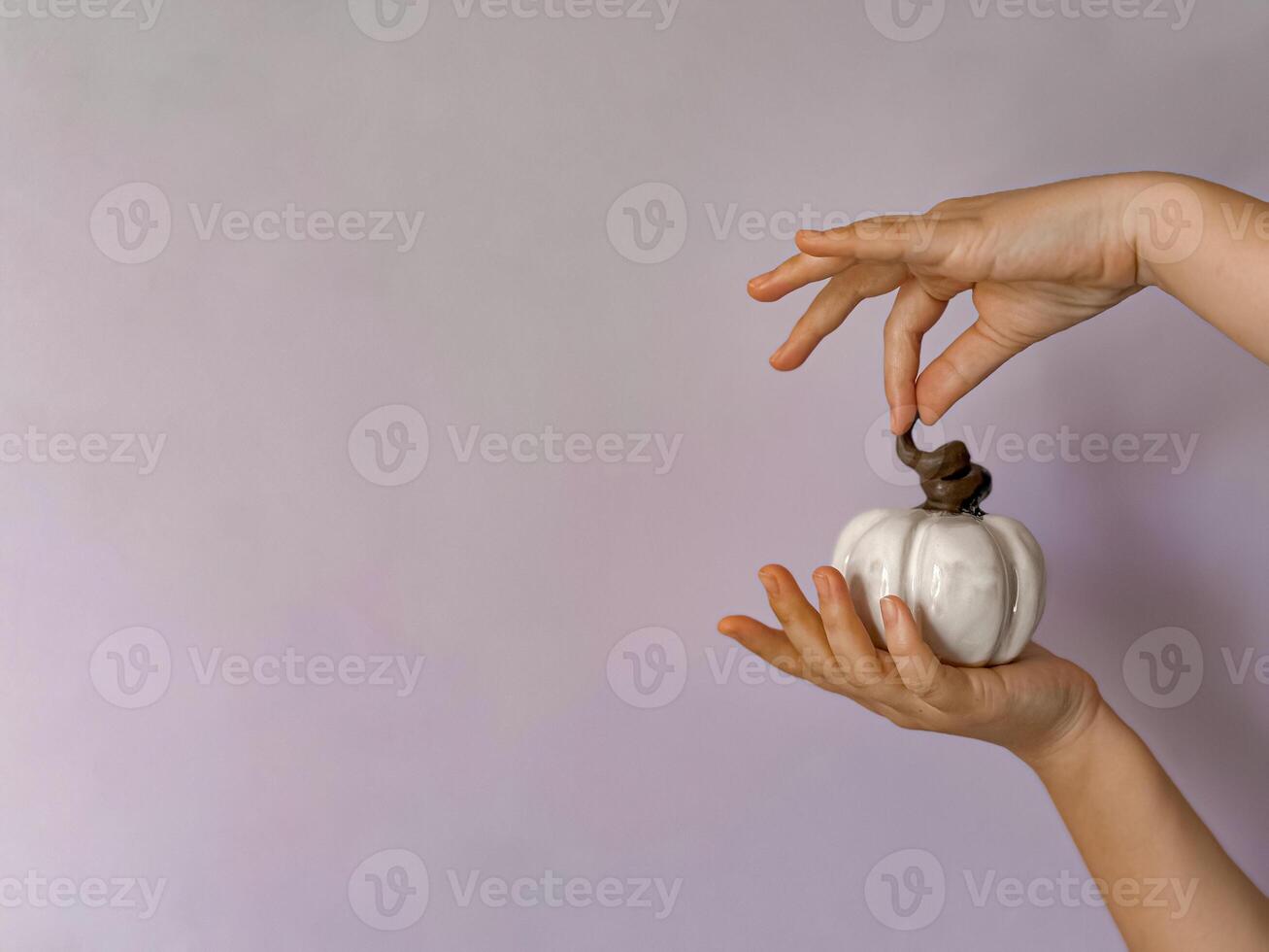 Close up of hand holding small white ceramic pumpkin with twisted brown stem against soft lavender background with copy space. Minimalist and symbolic representation of autumn, Halloween fall decor. photo