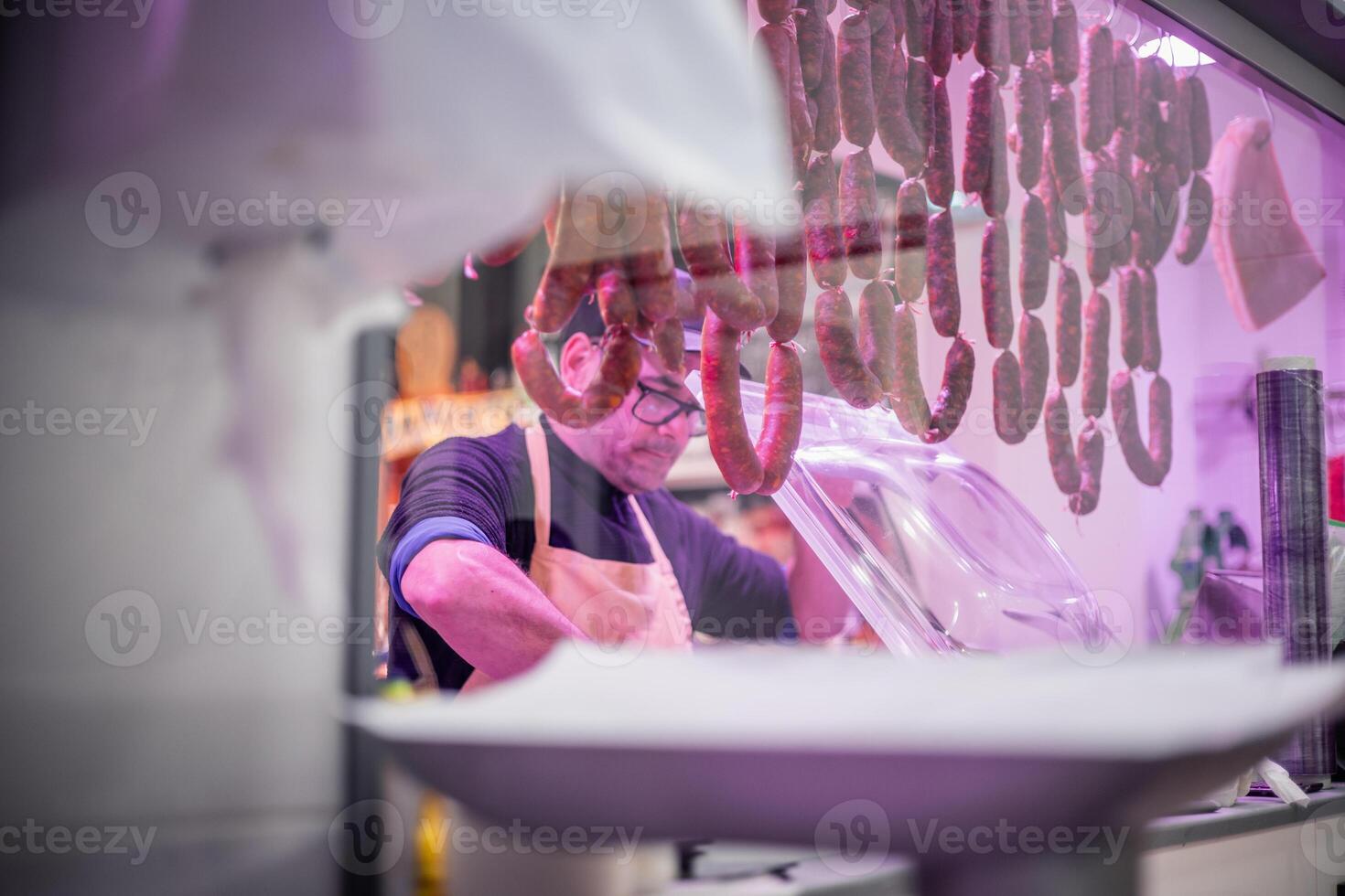 Butcher Using Vacuum Sealing Machine with Sausages and Chorizos Hanging from Butcher Shop Ceiling in Foreground photo