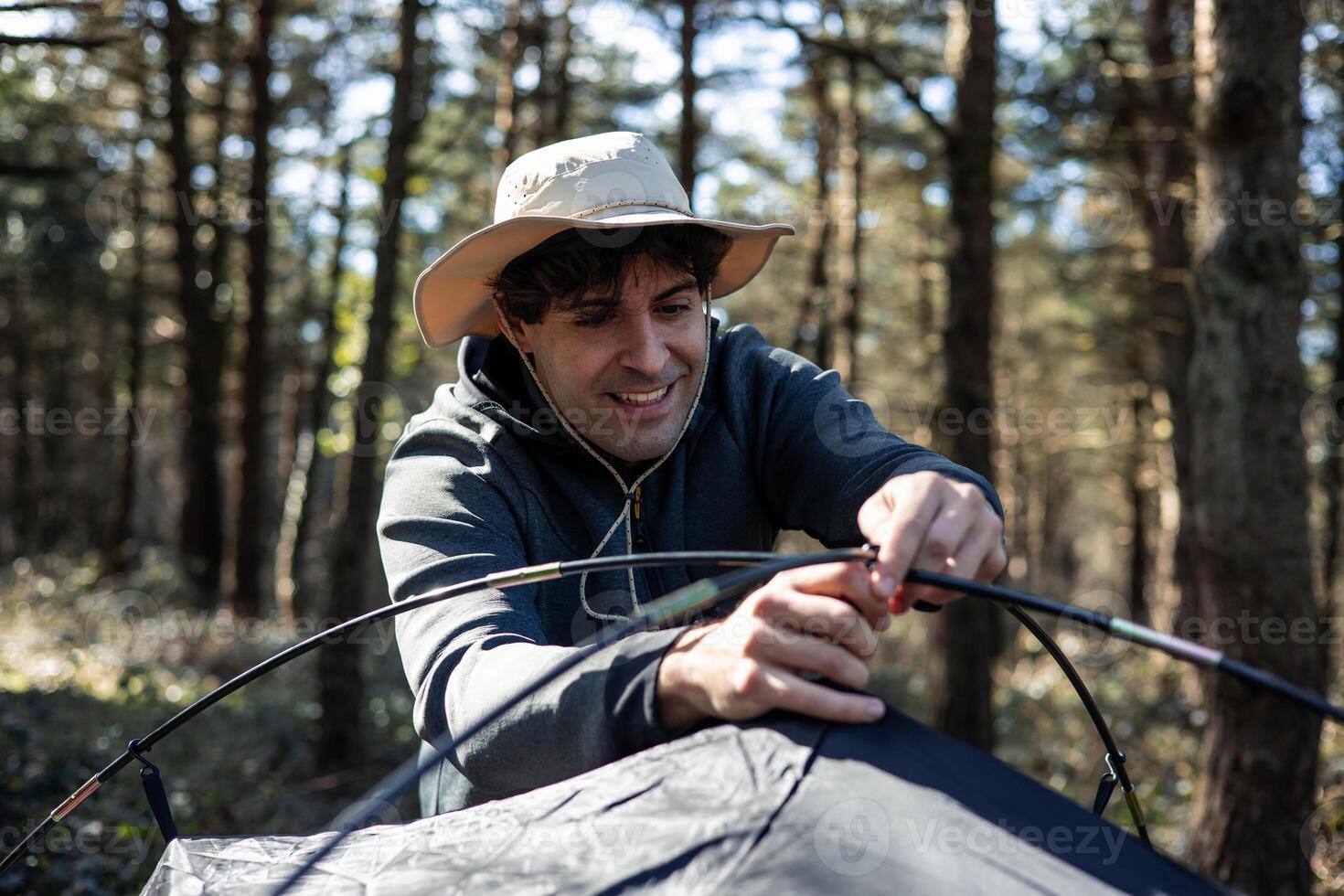 vertical portrait Smiling young man with hat assembling tent poles at campground during summer sunset photo