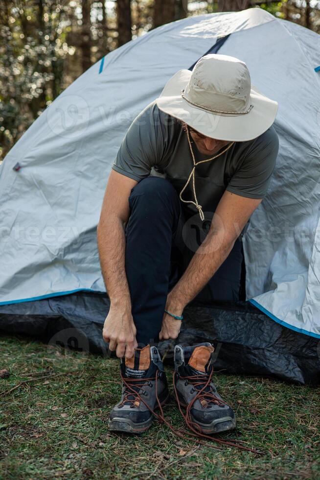 vertical portrait Explorer Camper Putting on Trekking Boots at Sunrise in Forest in front of his tent photo
