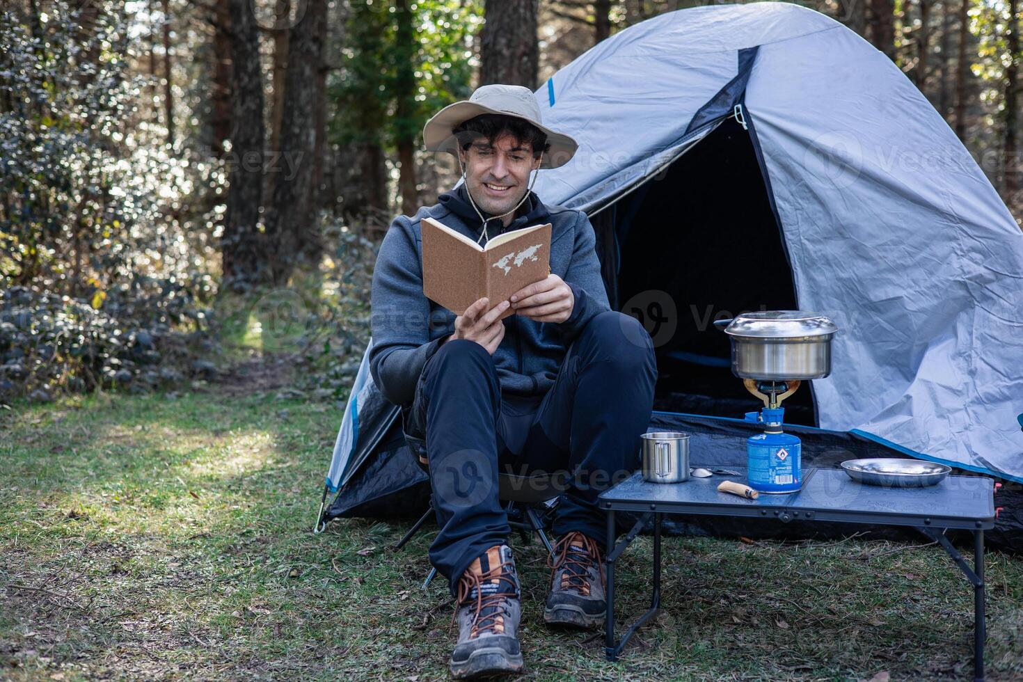 Man with explorer hat reading book at forest campsite while cooking on camping stove photo