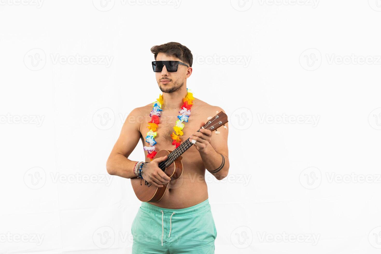 Young man in swim trunks playing ukulele with sunglasses and flower necklace on white background photo