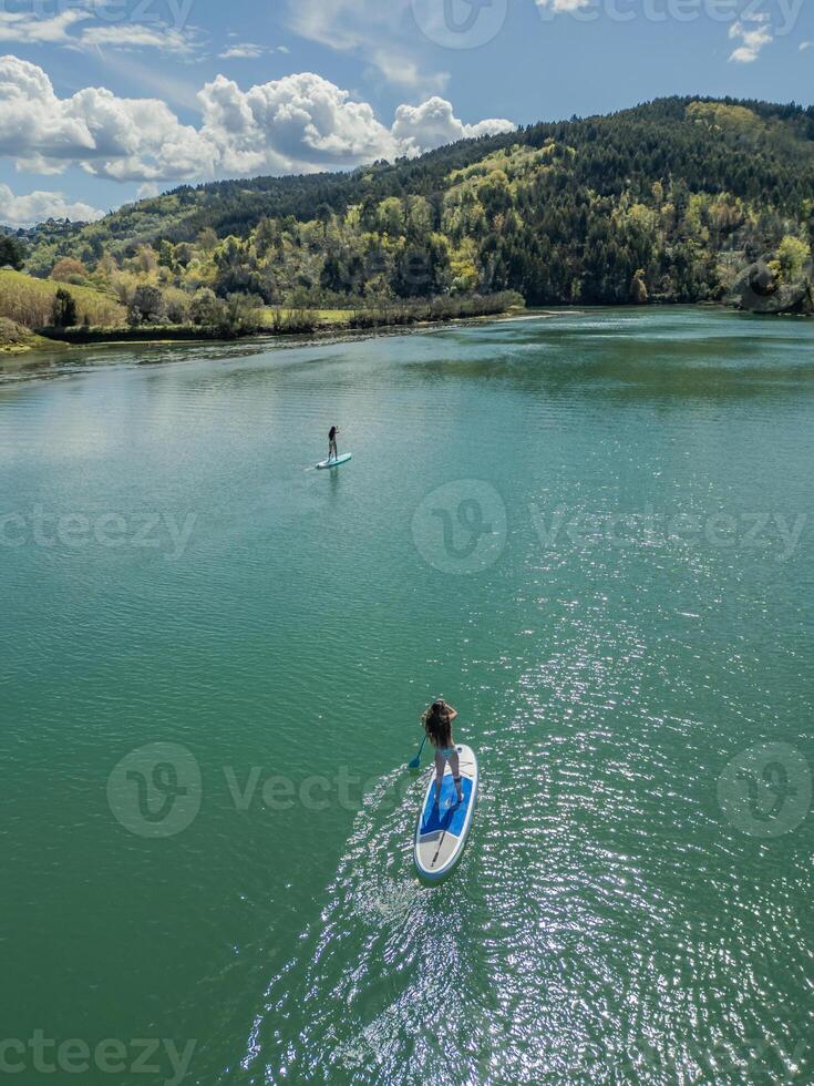 Two young girls paddleboarding on a river surrounded by nature on a sunny summer day photo