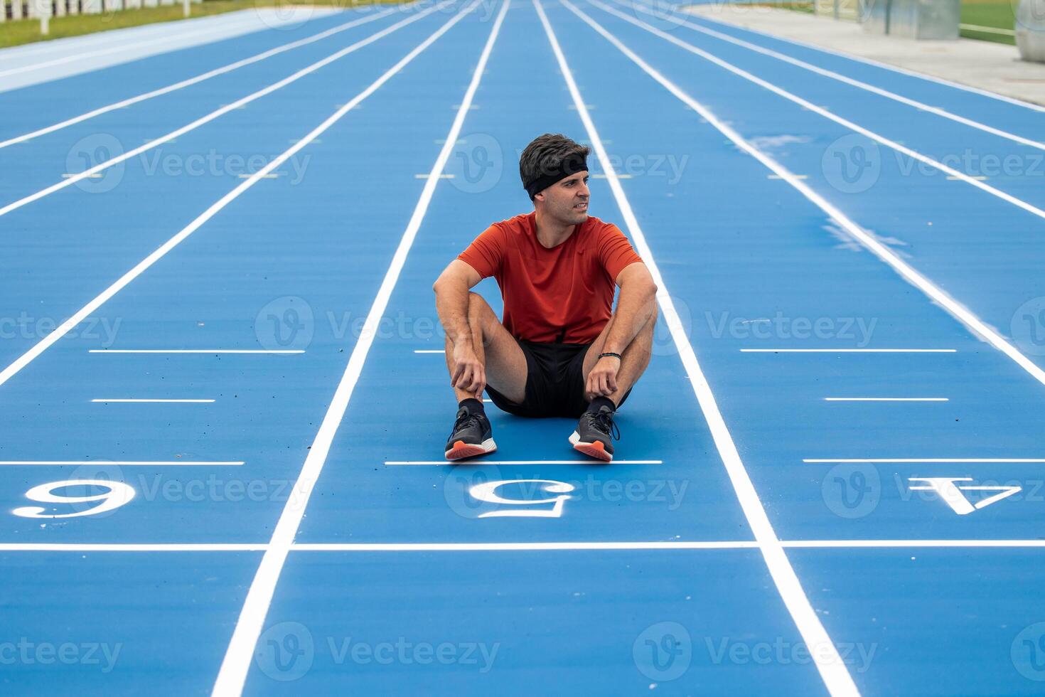 Male Runner with Headband Resting Seated on Finish Line of Blue Running Track photo
