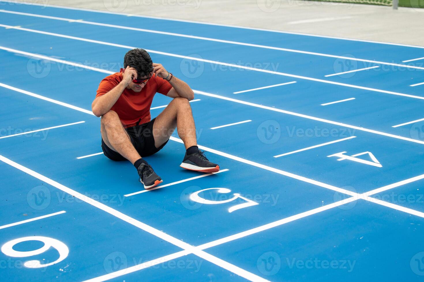 Exhausted Male Runner with Sunglasses Sitting on the Ground at Finish Line of Blue Running Track photo