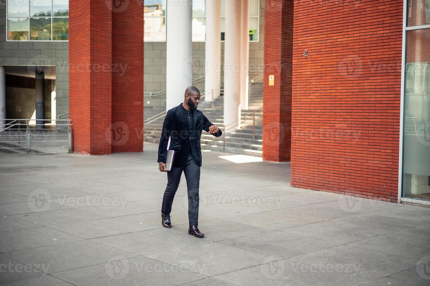 black African American businessman walking with laptop and checking watch in financial district photo