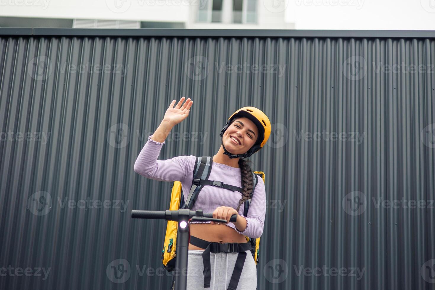food delivery girl on electric scooter with yellow backpack and helmet greeting with raised hand photo