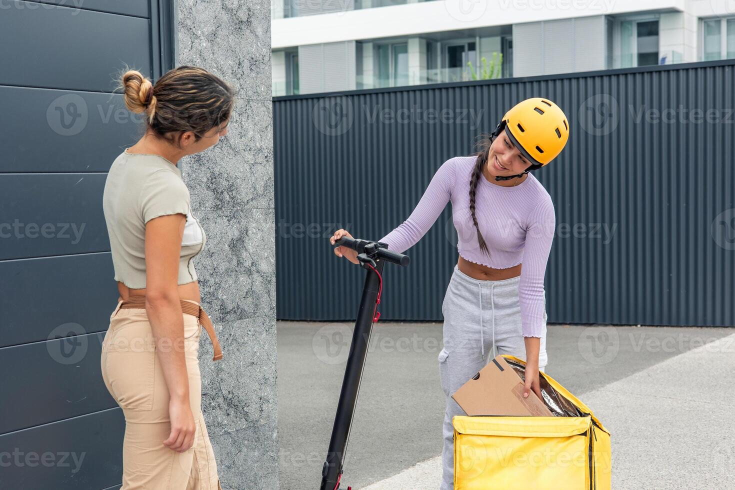 Latin courier with yellow helmet and electric scooter taking package from backpack for customer photo