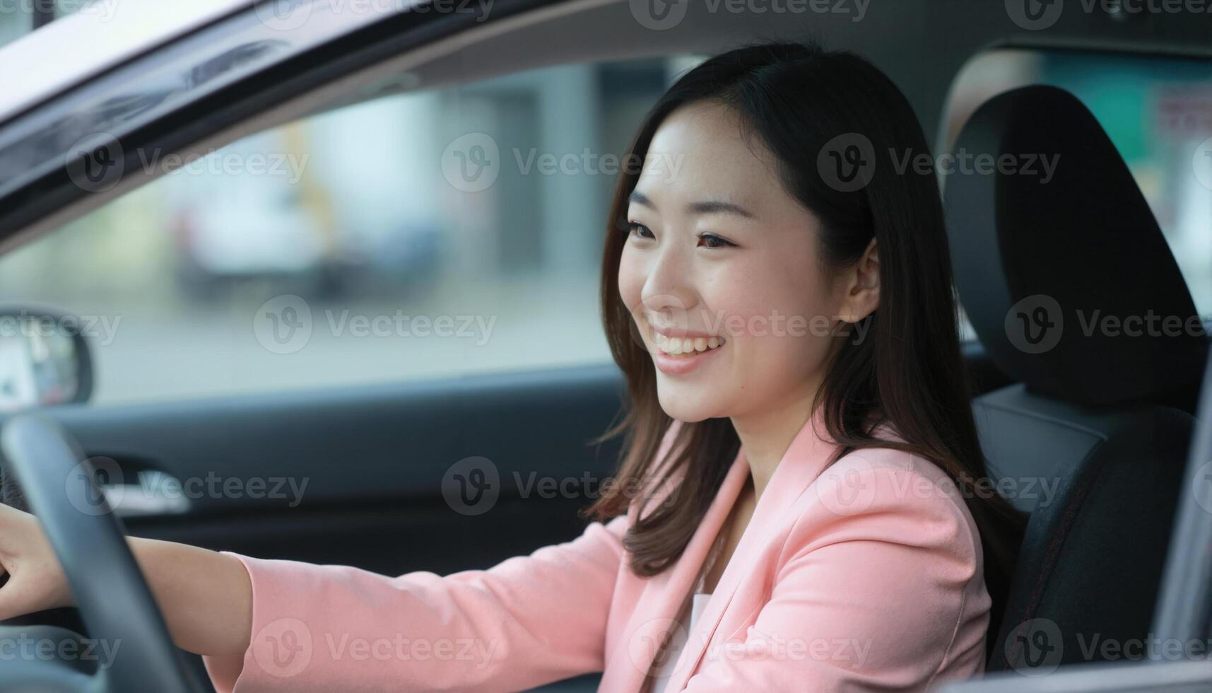 A smiling Asian woman in a pink blazer sits in the driver's seat of a car, holding the steering wheel. photo