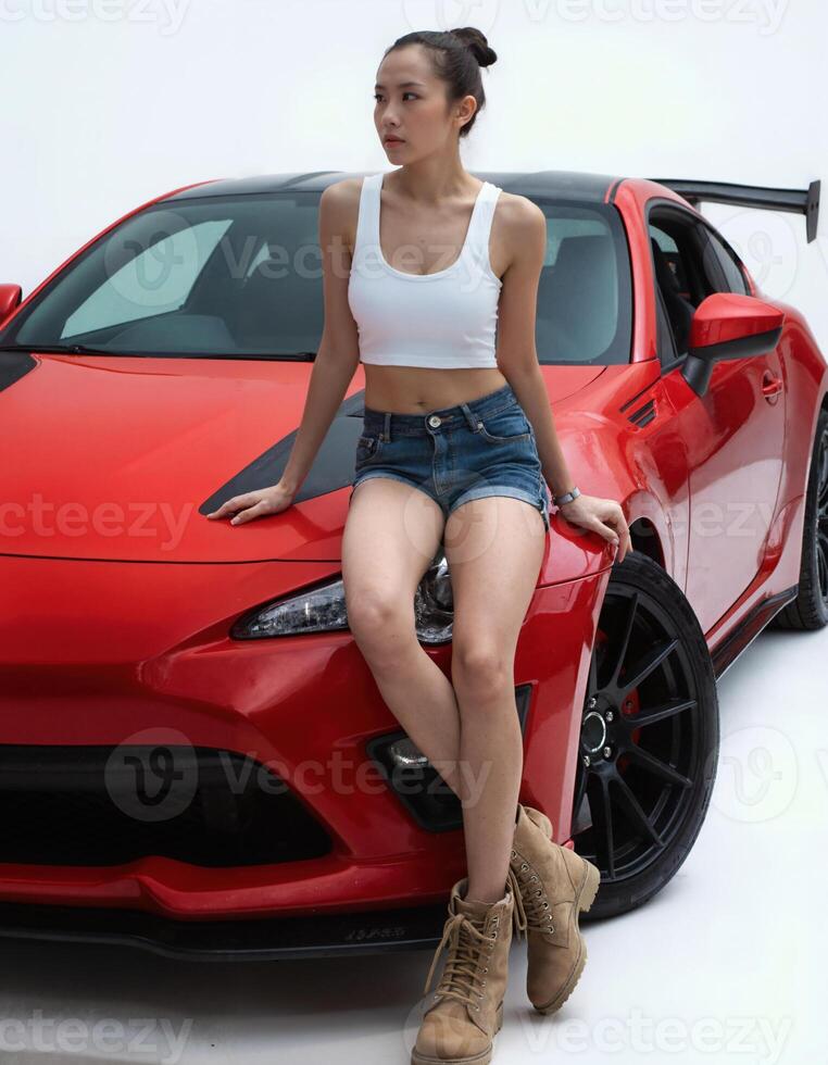 A young Asian woman with a slender physique, wearing a white crop top and denim shorts, sits on a red sports car. photo